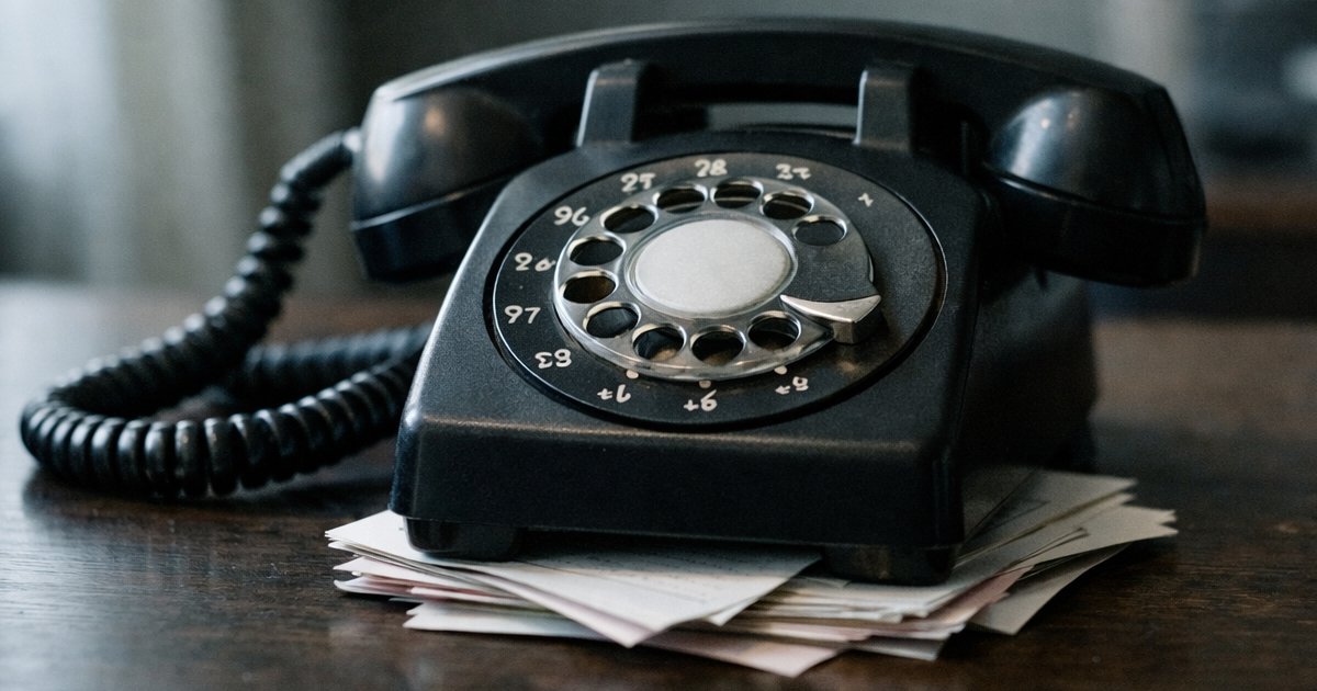 A close-up of a rotary office telephone sitting on a dark wooden desk. The receiver is slightly off the hook. A small stack of paper message slips accumulates underneath it. Soft, cool window light falls across the desk from the left. The room behind is blurred and dim. The mood is quiet neglect — not dramatic, just still. Shallow depth of field. Documentary photography aesthetic.